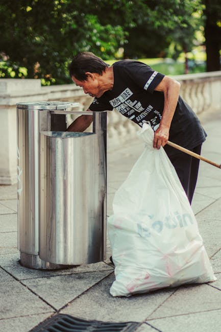 A man with dark hair, wearing a black T-shirt with white printed text on the front, is bending over a stainless steel recycling bin on a paved walkway surrounded by green trees and foliage. He is placing a large white plastic bag filled with waste into the bin, holding it open with one hand while the other hand stabilizes the bag. The waste bag appears to contain discarded items, possibly rubbish collected during street cleanup or waste disposal. The stainless steel bin has a cylindrical shape with a slot opening at the top for inserting waste, and it is positioned near a decorative stone railing in what appears to be an outdoor park or urban area. The scene is well-lit with natural daylight, emphasizing a clean environment and focusing on proper waste collection, consistent with the services provided by Rubbish Removal Lambeth for efficient waste management and alternative rubbish disposal options.