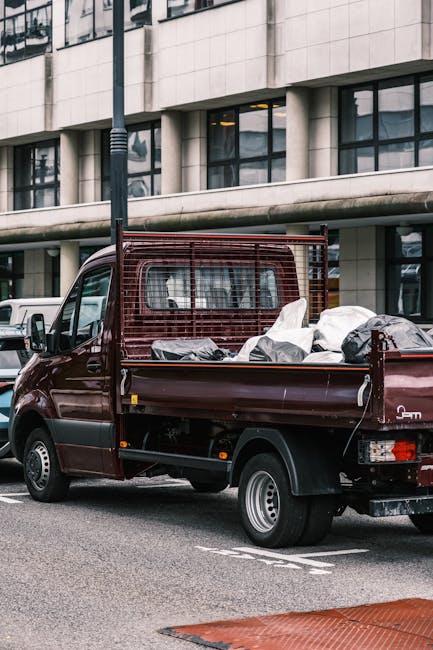 A small burgundy flatbed truck parked on an urban street in front of a modern office building with large glass windows and concrete exterior, loaded with various types of waste including several white and grey bags, some wrapped in plastic, and loose debris. The truck features a protective metal grid behind the cab, and its bed appears partially filled with rubbish ready for collection, suggesting an on-site clearance or private waste removal service. The environment is illuminated by natural daylight, with the surroundings including a streetlamp and parked cars, reflecting a typical city scene where independent rubbish removal companies, like Rubbish Removal Lambeth, might operate outside of local authority collection schedules for alternative waste handling.