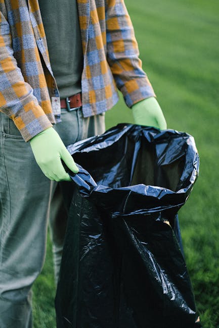 A middle-aged woman with short dark hair, wearing a black t-shirt with white text and dark trousers, is seen standing on a paved outdoor walkway next to a metallic, cylindrical rubbish bin with multiple sections for waste separation. She is holding a large white plastic bag in one hand and leaning forward with her other hand to dispose of rubbish into the bin. The surrounding environment features a low stone wall in the background, lush green trees, and sunlight filtering through the foliage, creating a bright and natural setting. The scene appears to be part of a public outdoor space, where individuals are engaged in waste disposal, reflecting a practical approach to rubbish removal and waste management outside of official collection services. The overall atmosphere is calm and orderly, indicating a responsible attitude towards waste disposal and environmental cleanliness, aligning with independent collection or on-site clearance practices that might be offered by rubbish removal services such as Rubbish Removal Lambeth.
