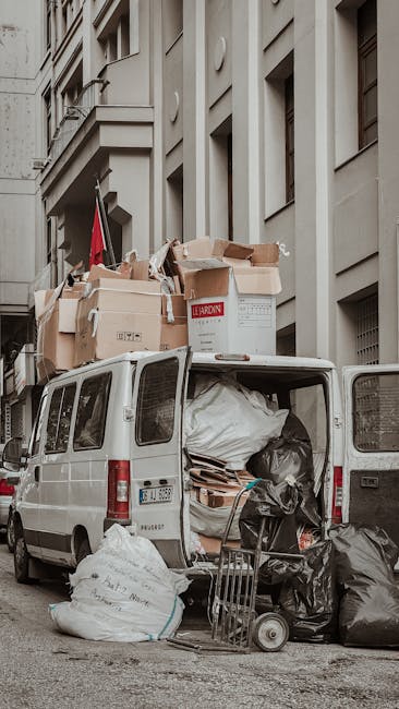A white commercial van parked on a city street adjacent to a multi-story building with light grey, textured facade and several rectangular windows. The back doors of the van are open, revealing a large load of mixed waste materials including black and white plastic bags, flattened cardboard boxes, and loose paper or packaging stacked unevenly inside. On top of the vehicle, there are additional cardboard boxes, some with printed labels, and a red flag attached to the building near the van. Outside the van on the pavement, there are more waste bags, and a small hand trolley or dolly leaning against the open rear doors. The scene suggests an on-site waste collection or clearance, with a focus on bulk rubbish removal, ideally handled by local private waste disposal providers such as Rubbish Removal Lambeth, facilitating alternative waste handling solutions for household or commercial refuse.
