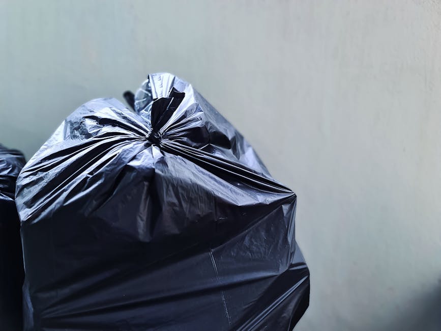 Close-up view of a tightly tied black plastic rubbish bag with a crinkled texture, placed against a plain, light-colored wall, suggesting indoor or outdoor waste collection activity. The bag appears full and is situated in the foreground, with additional bags partially visible behind it, indicating a collection of household waste or refuse awaiting removal. The environment seems clean and free of clutter, with no other objects or environmental details visible. The focus on the black trash bag aligns with private rubbish disposal services such as those offered by Rubbish Removal Lambeth, highlighting materials typically handled during on-site clearance or alternative waste handling efforts. The neutral lighting emphasizes the shiny, reflective surface of the plastic, providing a clear and neutral visual suitable for accessibility and SEO relevance related to rubbish collection and waste management.