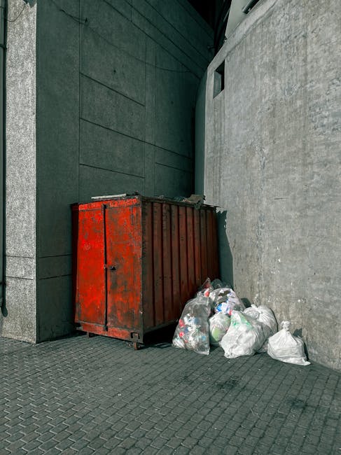 A red metal waste container with visible rust and a weathered surface is positioned against the corner of a building, with its front slightly ajar. Surrounding the container are several tightly tied white plastic rubbish bags filled with general household waste, some leaning against the container's side on a paved area. The background features the dark, smooth wall of a modern building with a textured grey concrete wall on the right, and a dark metallic or composite cladding surface to the left. The scene is illuminated by natural daylight, casting soft shadows on the ground. This outdoor setting, located on a paved urban space, illustrates a typical instance of independent rubbish disposal that could be handled by private waste removal services such as Rubbish Removal Lambeth, especially suitable as an alternative to council-managed bulky waste collection, provided the waste conforms to local regulations for disposal.