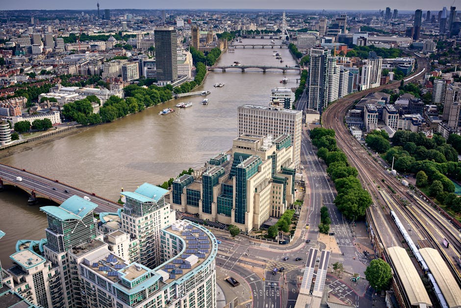An aerial view of central London showing the River Thames flowing through the city with several boats visible on the water. On the southwest bank, there are modern office buildings with distinctive glass facades, some featuring blue-tinted windows and rooftop terraces. To the right, a railway track runs parallel to a street lined with evenly spaced green-leafed trees, with a train station and station platforms visible. The surrounding area includes a mix of high-rise towers, mid-rise offices, and residential buildings, creating a dense urban landscape. Bridges crossing the Thames are visible in the background, connecting different parts of the city. The scene is well-lit with natural daylight, highlighting the varied textures of glass, concrete, and steel in the cityscape. The image contextually relates to city infrastructure and urban development, aligning with topics such as urban planning and the importance of efficient waste management in metropolitan areas, as well as the role of independent rubbish removal services like Rubbish Removal Lambeth in maintaining city environments.