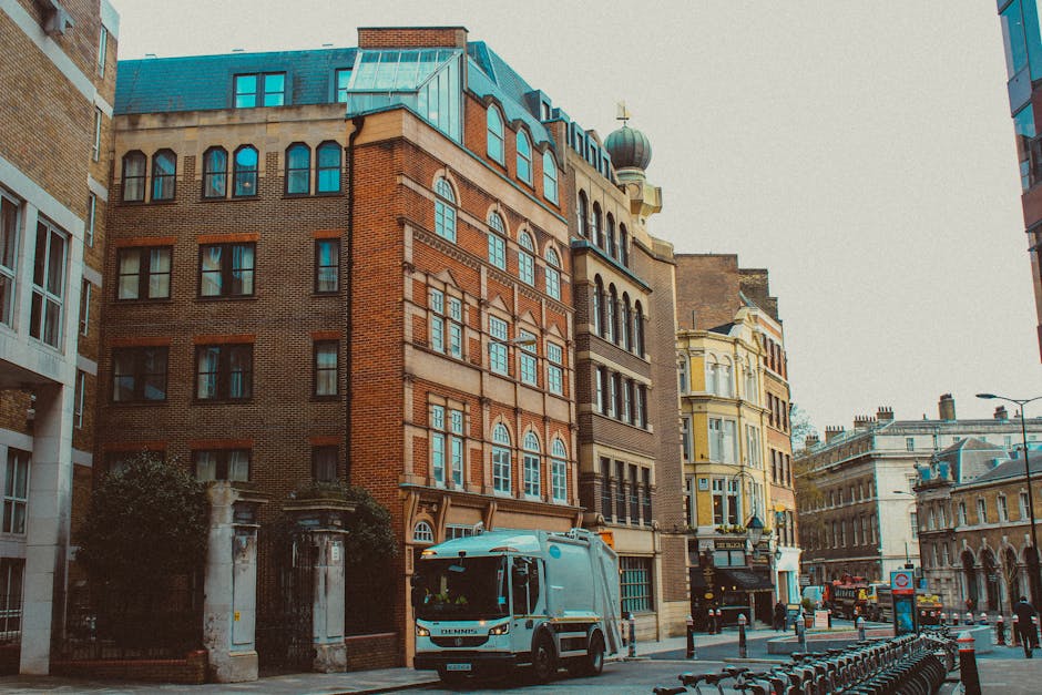 A street view featuring a mix of historic and modern buildings with brick and stone facades, large windows with arched and rectangular frames, and decorative architectural details. In the foreground, there is a light blue waste collection vehicle parked on the paved sidewalk, which is lined with metal bollards and bike racks. To the left, a gated entrance with stone pillars and shrubbery surrounds a small property, while further along the street, additional vehicles and street lamps are visible against a grey, overcast sky. The environment appears urban with a focus on transport and street activity, aligning with private waste collection or rubbish removal services. Rubbish Removal Lambeth occasionally supports such on-site clearance efforts through their comprehensive rubbish services, as suggested by the street scene contextual relevance. The overall scene emphasizes the typical street scene in central London, with a subdued atmosphere that complements the theme of rubbish removal and urban waste management.