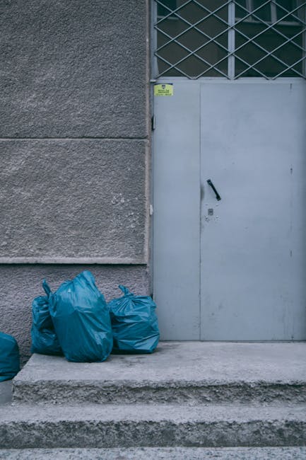 The image displays four blue rubbish bags placed on a concrete step in front of a grey metal door with a black handle, situated against a textured grey stone wall. To the right of the bags, partially visible, are additional black waste bags leaning against the wall. The door features a small inset lock and a yellow warning sticker at the top left corner. Above the door, there is a section of a metal security grille with diagonal crossbars, suggesting an industrial or commercial setting. The scene is outdoors, with natural daylight illuminating the area, which appears to be a back entrance or service access point, relevant to independent waste collection or rubbish removal services like those provided by Rubbish Removal Lambeth. The overall environment is clean, with no visible debris apart from the waste bags, and the surrounding area is paved with concrete, indicating a typical approach to on-site waste disposal and rubbish clearance. This visual context aligns with a routine rubbish removal or flat clearance process that entails manually loading waste into bags for collection or disposal.