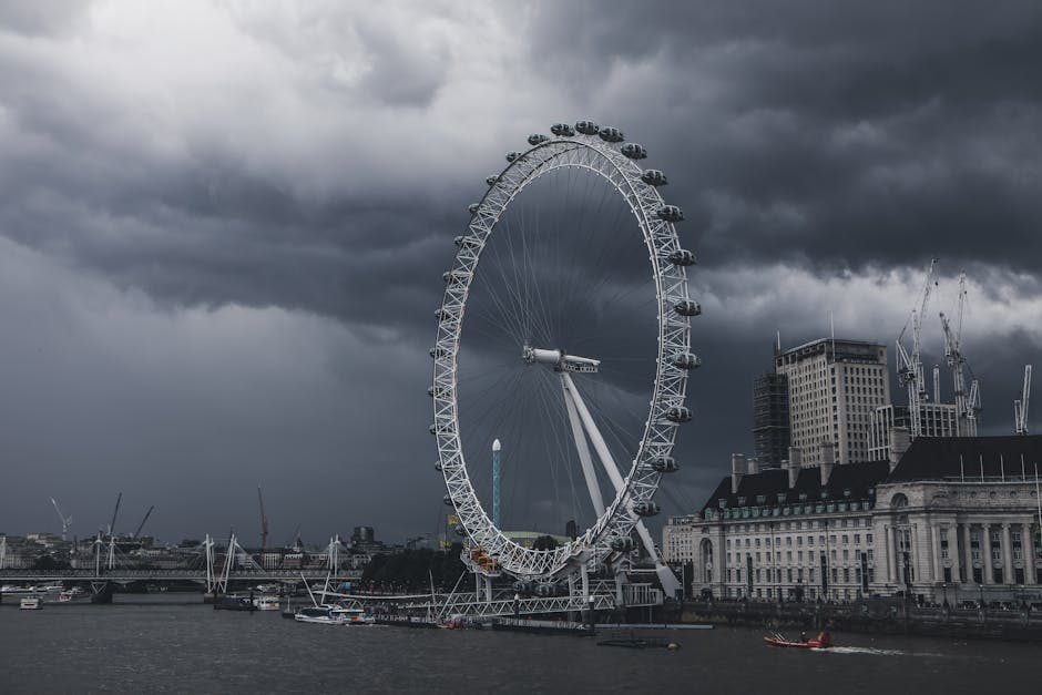 A large Ferris wheel with multiple enclosed passenger capsules is situated along the waterfront, with a cloudy, dark sky overhead indicating an impending storm. The Ferris wheel's white metal structure contrasts with the heavy, dark gray clouds in the background. To the right of the wheel, there are several mid-rise buildings with a mix of modern and historic architectural styles, some featuring cranes indicating ongoing construction. In the foreground, boats and small vessels are docked along the water's edge, which appears to be part of a busy urban harbor. The overall scene is characterized by a moody atmosphere with subdued lighting, emphasizing the scale of the Ferris wheel and the surrounding cityscape, relevant to the context of managing large-scale outdoor structures and urban environments in waste and rubbish removal considerations, as handled by services like Rubbish Removal Lambeth.