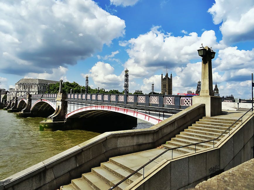 A wide-angle view of Westminster Bridge spanning the River Thames in London under a partly cloudy sky. The bridge’s decorative metal railings are painted in a light pinkish hue with darker accents, and the stone pillars at each end feature ornate lamp posts. The river below shows rippling water with greenish-brown tones. In the background, the tall, historic clock tower known as Big Ben rises above surrounding buildings, including Parliament, with their Gothic architectural details visible. The foreground includes a stone staircase with a metal handrail leading down to a walkway adjacent to the riverbank, which is clean and well-maintained. The scene suggests an urban environment with a mix of classical and early 20th-century architecture, capturing the iconic London riverfront scene that could relate to private or independent disposal options near famous landmarks, illustrating the city’s blend of historic structures and modern infrastructure, relevant to waste management and rubbish removal services in central London.
