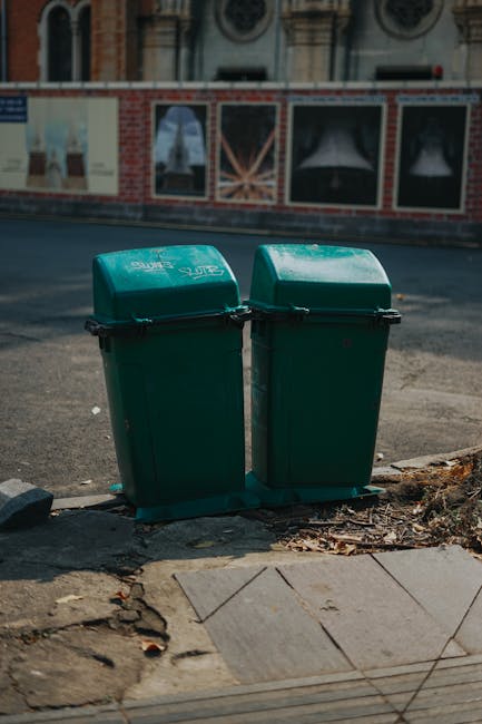 Two green wheeled rubbish bins with hinged lids positioned side by side on a sidewalk at the edge of a paved area, close to a curb, in front of a building with a brick facade and large display windows. The bins are made of durable plastic with a smooth, slightly reflective surface and black handles securing the lids. Surrounding the bases of the bins, there are small scattered leaves and dirt, indicating an outdoor setting in an urban environment. The pavement consists of concrete slabs with visible joints, and the curb is made of rougher, darker material. The background shows parts of the street and the lower sections of a building’s window displays, suggesting a typical commercial or residential area. The scene is naturally lit, with daylight casting slight shadows, and the presence of the bins hints at private waste collection or alternative rubbish disposal services, such as those provided by Rubbish Removal Lambeth for local rubbish management near Lambeth Palace.