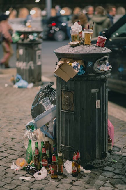 A black, cylindrical metal rubbish bin with a domed lid situated on a cobbled pavement in an urban outdoor setting. The bin is surrounded by scattered trash, including several empty glass bottles, crumpled paper, and plastic packaging located at its base, with some debris leaning against its side. The top of the bin holds a pile of discarded food and drink containers, such as a partially crushed cardboard food container, two takeaway cups with plastic lids and straws, and a small plastic cup, all appearing to be used. Additionally, there are some paper bags and wrapping materials placed on the lid, with a few items visibly spilling over the edges. In the background, a grey public trash bin and a group of blurred pedestrians are visible, along with parked cars and some out-of-focus cityscape elements. The scene captures a typical urban waste presentation, aligning with private waste handling or alternative rubbish collection services like those offered by Rubbish Removal Lambeth.