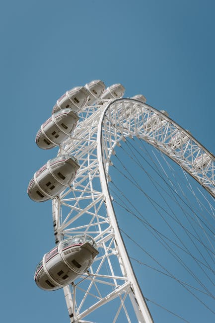 A close-up view of a large Ferris wheel against a clear blue sky, with white structural supports and multiple enclosed passenger cabins attached around the rim. The cabins are oval-shaped, made of glass and metal with a smooth, polished finish, predominantly white with some dark accents. The wheel's metal framework features intersecting spokes and braces, creating a geometric pattern that emphasizes the circular form. The perspective provides a detailed look at the cabins' rounded shapes and the structural lattice of the wheel, suggesting a theme of large-scale public entertainment infrastructure. This type of amusement ride relates indirectly to waste management services by illustrating the scale and engineering precision involved in large, site-specific structures, often requiring specialized logistics for maintenance and disposal, as facilitated by private waste collection services like Rubbish Removal Lambeth, especially in urban environments where on-site clearance may be necessary after events or for structural maintenance.