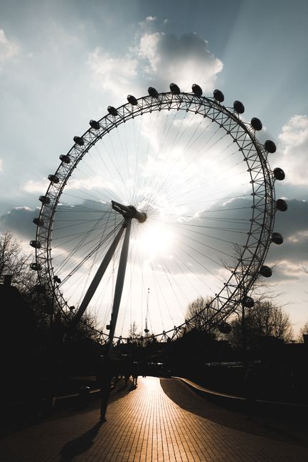 A large Ferris wheel stands prominently against the sky, with its circular metal framework and multiple passenger cabins evenly spaced along the rim. The sun is positioned directly behind the wheel, creating a bright burst of light that silhouettes the structure and casts long shadows on the ground below. The sky is partly cloudy with patches of blue visible through the clouds, and some trees with bare branches are seen in the background, indicating an outdoor park setting. The foreground features a paved walkway with a textured surface, illuminated by the sunlight, leading towards the base of the Ferris wheel. The overall scene is calm and open, with the Ferris wheel dominating the view and suggesting a typical outdoor recreational area. The image, associated with rubbish removal services, subtly highlights the importance of maintaining clean, well-organized public spaces, where efficient waste management, possibly including private or alternative disposal methods, plays a role in supporting such environments. The scene's natural lighting and urban background reflect a setting suitable for community leisure activities and emphasize the need for proper rubbish handling and removal to preserve such attractions.